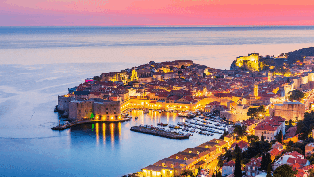 Illuminated Dubrovnik harbor and historic city walls overlooking the Adriatic Sea at sunset.